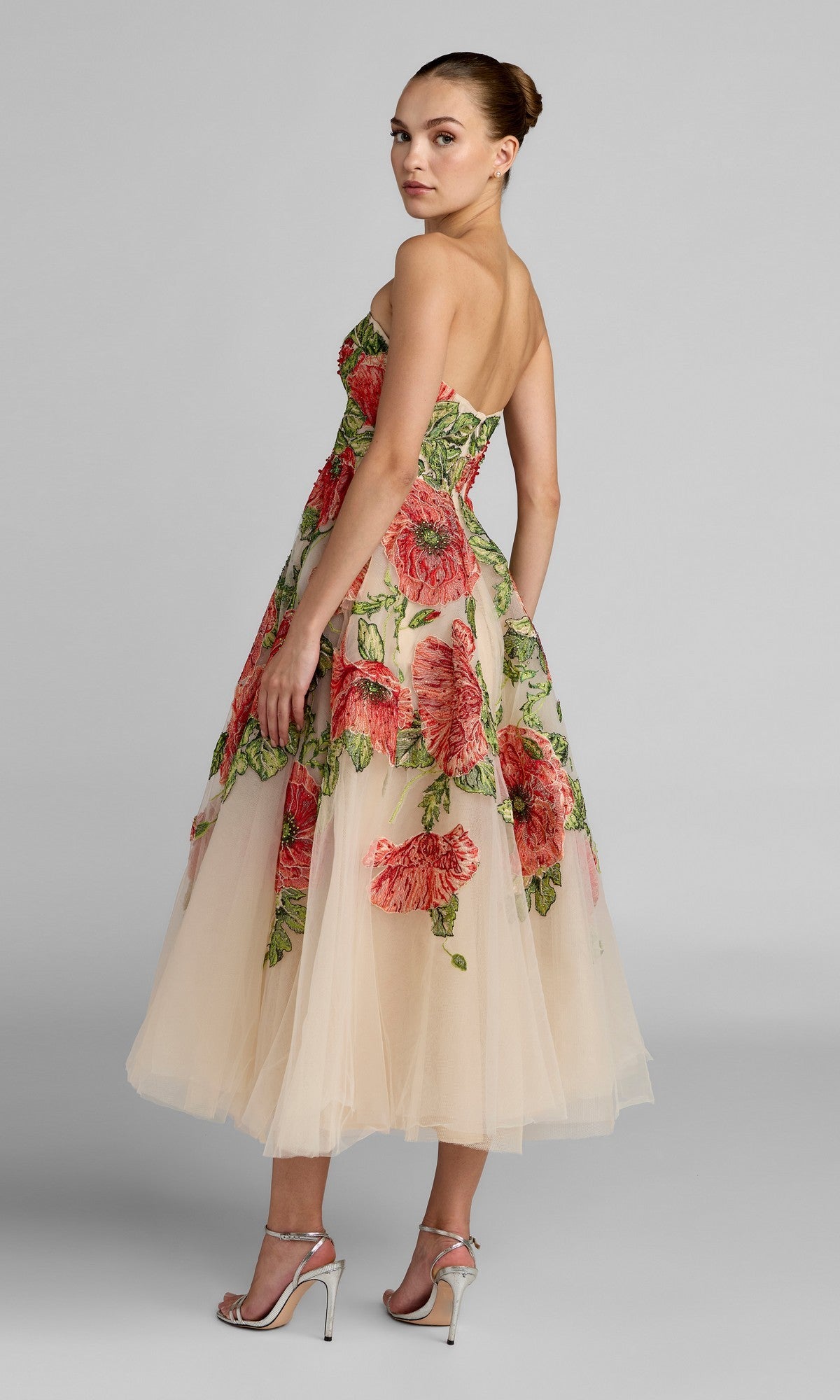 A woman stands sideways, looking over her shoulder, dressed in the Tea-Length Party Dress: Mac Duggal 21300—a strapless tulle midi dress adorned with bold red floral embroidery. She completes the sophisticated look with silver high-heeled sandals, all set against a gray background.